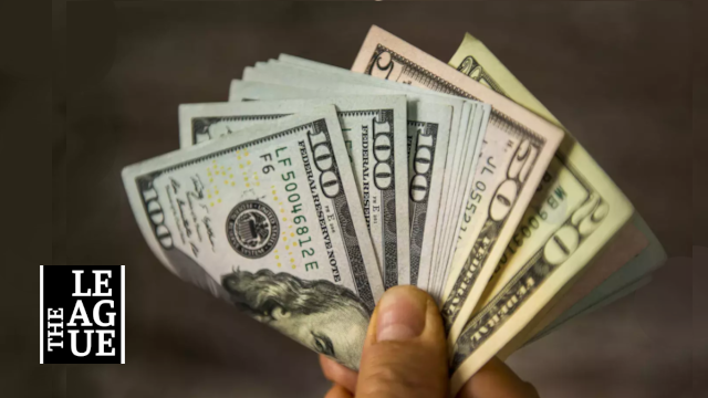 Close-up of a hand fanning multiple US dollar bills (various denominations) against a dark background, with a logo in the bottom-left corner.